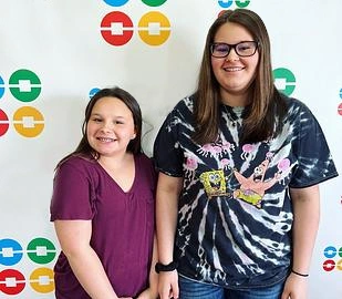 Two smiling girls standing together at orthodontics event with colorful logo backdrop.
