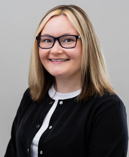 Professional headshot of smiling woman wearing glasses and black cardigan against neutral gray background.