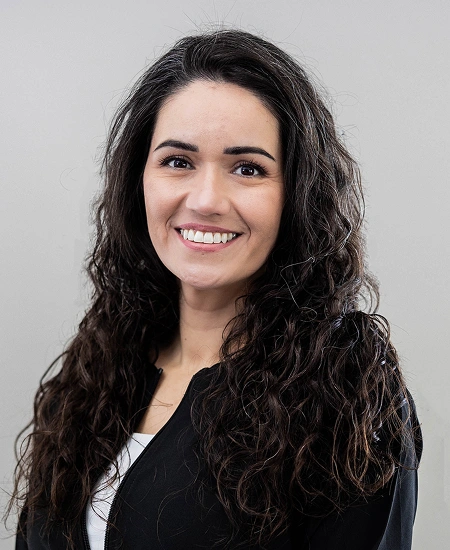 Professional headshot of smiling woman with long dark curly hair wearing black blazer against gray background.
