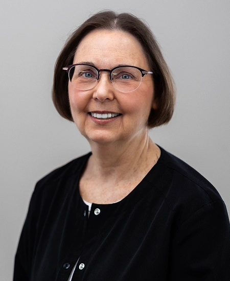 Professional headshot of a woman with glasses and dark hair wearing a black cardigan against a gray background.