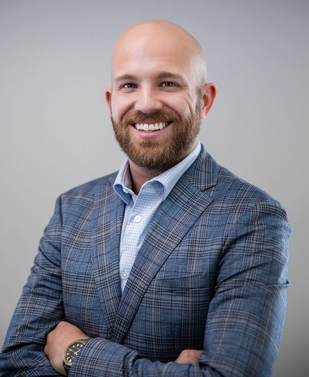 Professional headshot of smiling man in blue plaid blazer against gray background.