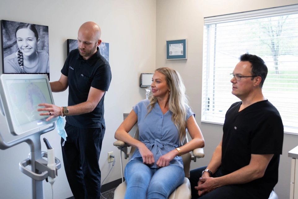 Orthodontist demonstrates dental equipment to seated female patient while male colleague observes in clinical office setting.